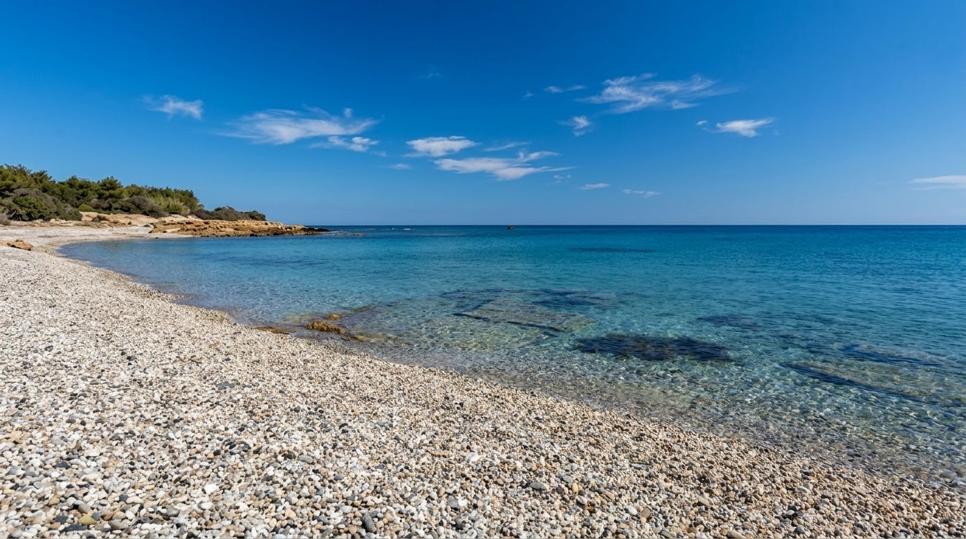 Porto Corallino, Villaputzu — spiaggia di ciottoli chiari con mare turchese trasparente, fondali rocciosi affioranti e macchia mediterranea sul promontorio