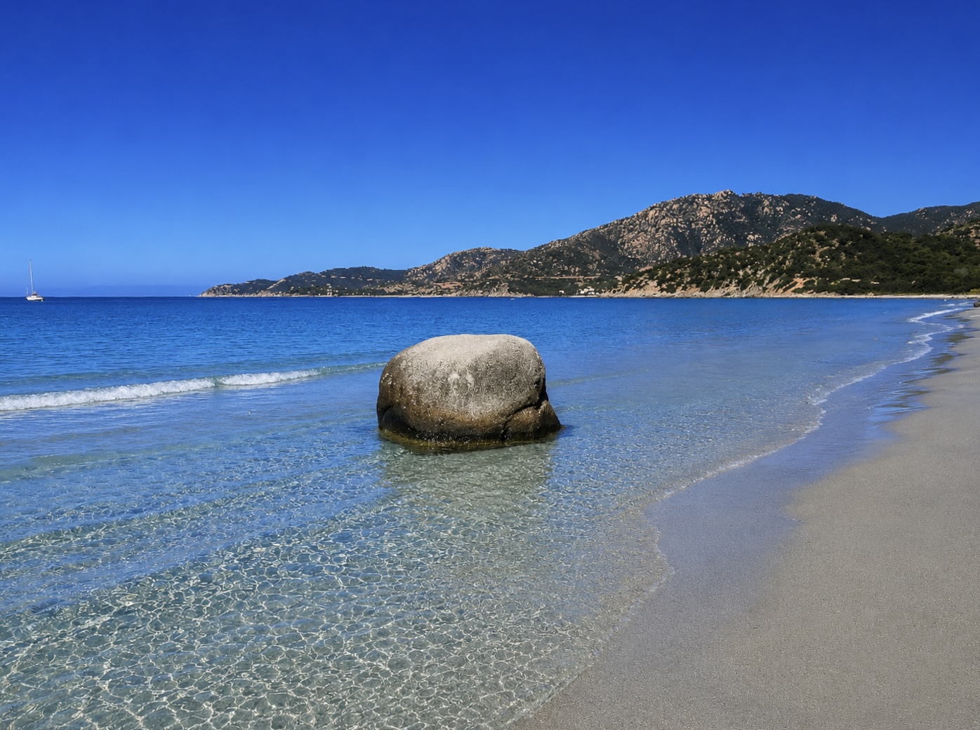 Campulongu, Villasimius — sabbia chiara con scogli bassi e acqua cristallina turchese, barche in rada e monti sullo sfondo
