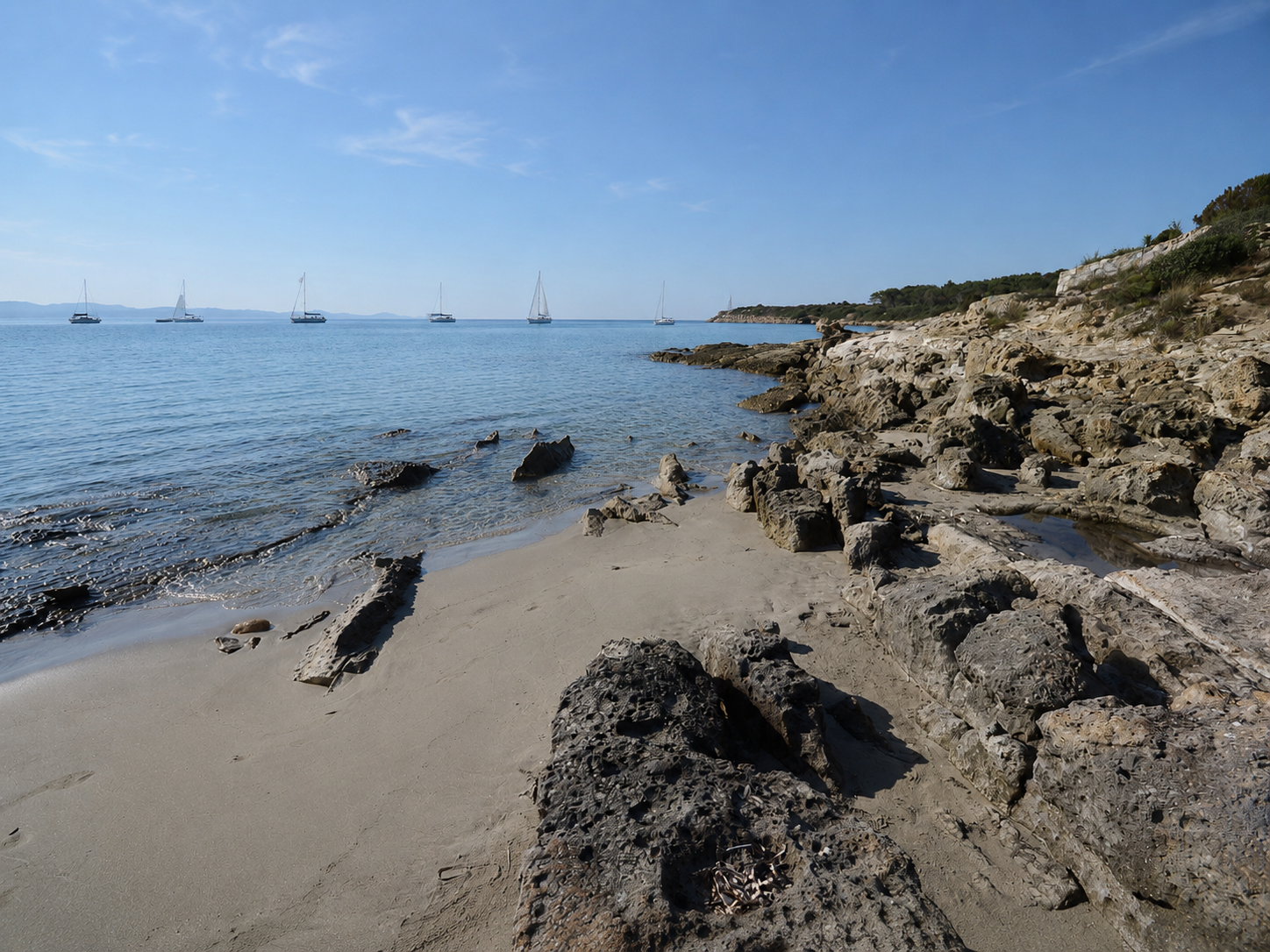 Guidi, Carloforte — piccola caletta di sabbia chiara tra scogli scuri con mare calmo e vele all'orizzonte
