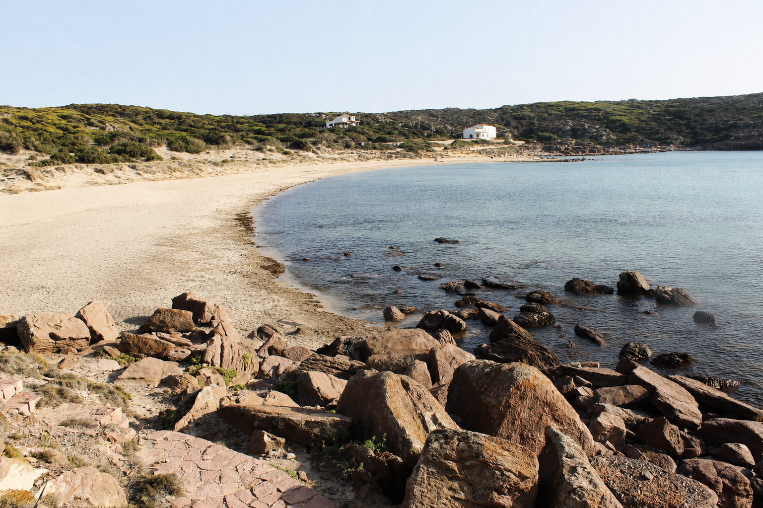 La Caletta, Carloforte — baia di sabbia chiara con scogli rossastri, dune verdi e acqua calma dell'isola di San Pietro