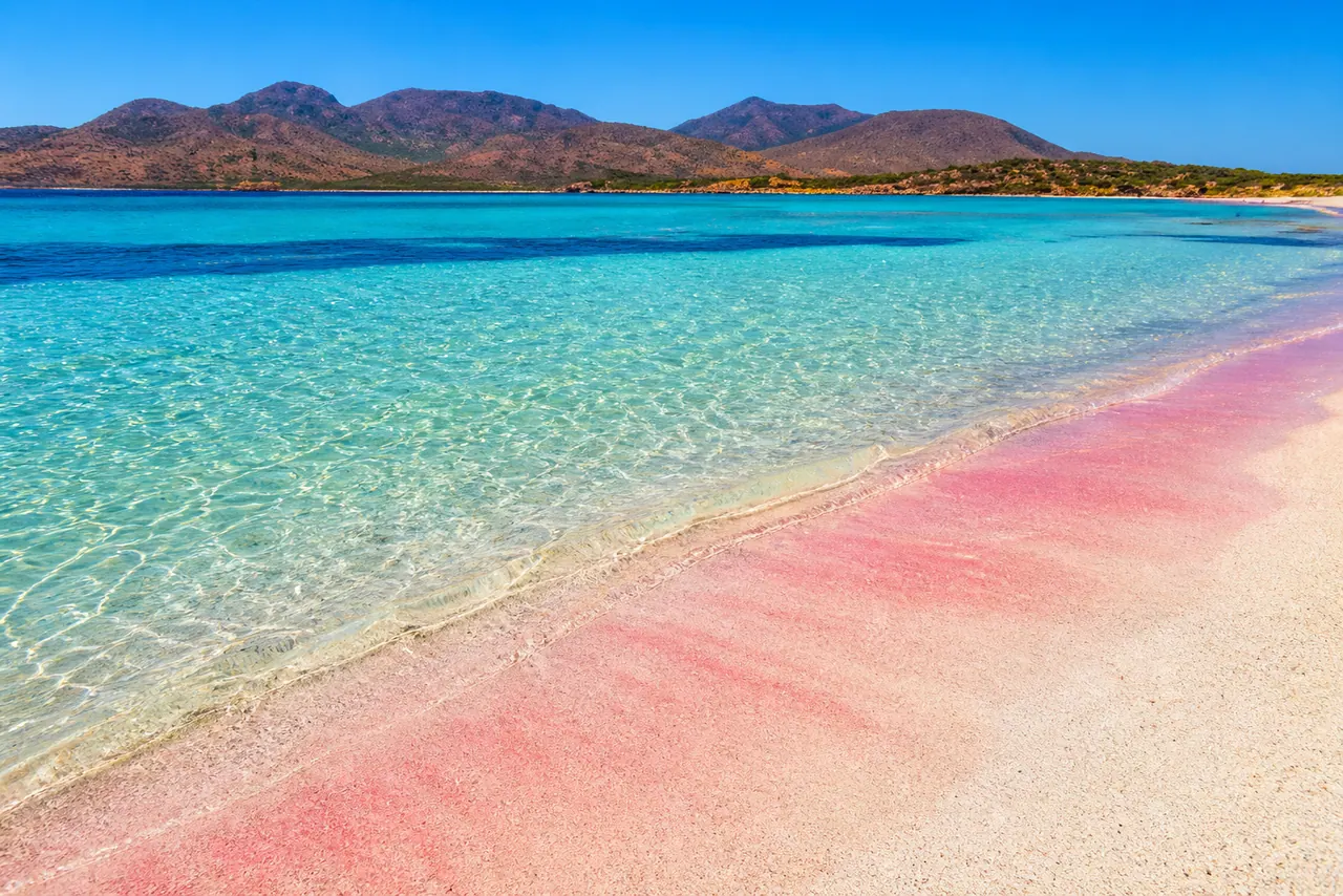 Cala Zafferano, Teulada — spiaggia di sabbia rosa con acqua turchese cristallina e monti del poligono sullo sfondo