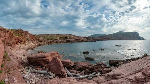 Cala del Gallo, Alghero — cala rocciosa con arenarie rosse, ciottoli e legni sulla riva, mare calmo e promontori all'orizzonte