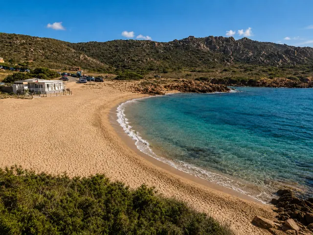 Cala Sarraina, Trinità d'Agultu — baia di sabbia dorata con acqua turchese cristallina, scogliere granitiche rossastre e macchia mediterranea sulle colline