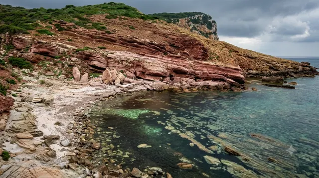 Cala Viola, Alghero — piccola cala di rocce rosse e ciottoli con acqua trasparente e cielo nuvoloso sul promontorio costiero