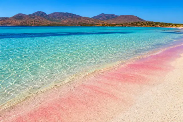 Cala Zafferano, Teulada — spiaggia di sabbia rosa con acqua turchese cristallina e monti del poligono sullo sfondo