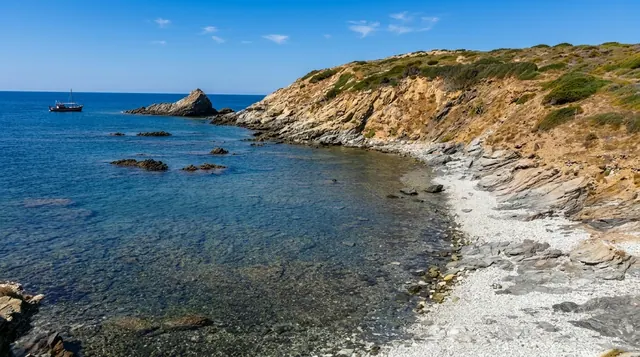 Coscia di Donna, Stintino — piccola cala di ciottoli bianchi tra scogliere stratificate, acqua trasparente e barca a vela al largo