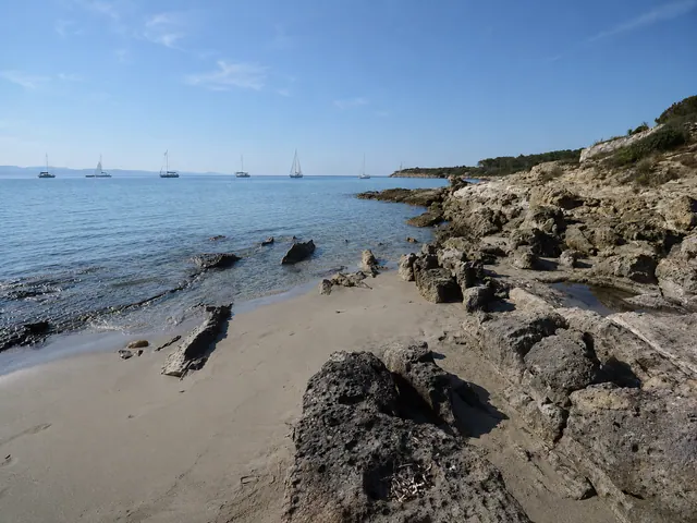 Guidi, Carloforte — piccola caletta di sabbia chiara tra scogli scuri con mare calmo e vele all'orizzonte