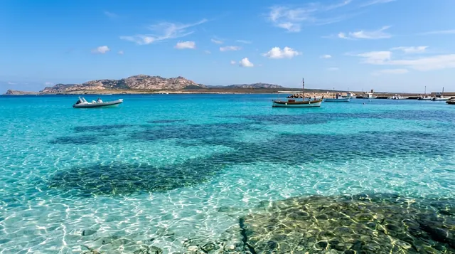 L'Ancora, Stintino — acqua cristallina bassa con fondale visibile, barche ormeggiate e profilo dell'Asinara sullo sfondo