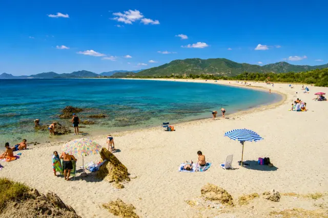 Porto Corallo, Villaputzu — ampia baia di sabbia chiara con mare blu, ombrelloni e profilo dei monti sullo sfondo
