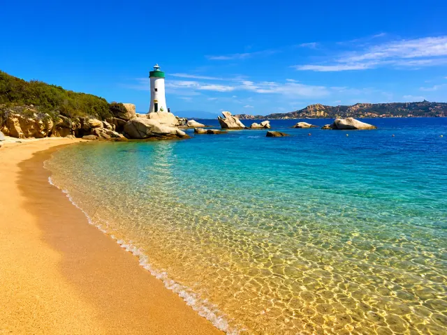 Porto Faro, Palau — spiaggia dorata con faro bianco sugli scogli e mare cristallino davanti all'arcipelago