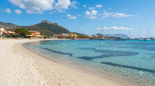 Prima Spiaggia, Golfo Aranci — baia urbana con sabbia chiara, acqua turchese, yacht ormeggiati e il profilo del promontorio di Capo Figari