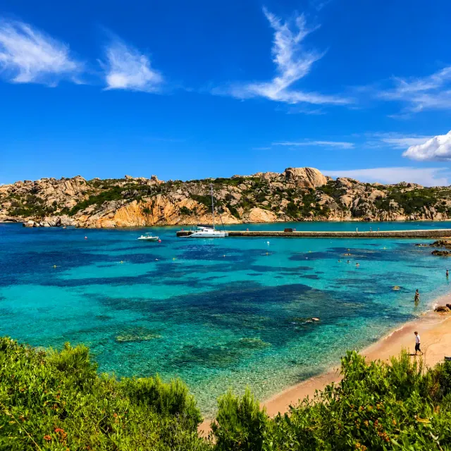 Cala Spalmatore, La Maddalena — baia turchese con pontile, scogli granitici e piccola spiaggia incorniciata dalla macchia
